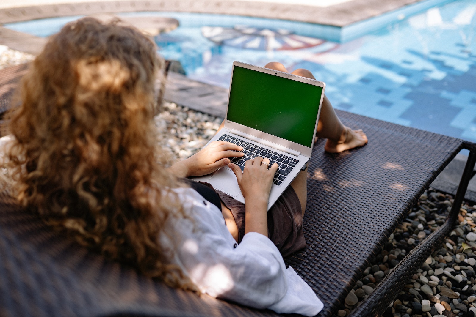 From above back view of anonymous female freelancer typing on portable computer while lying on sunbed near swimming pool with transparent water in sunlight